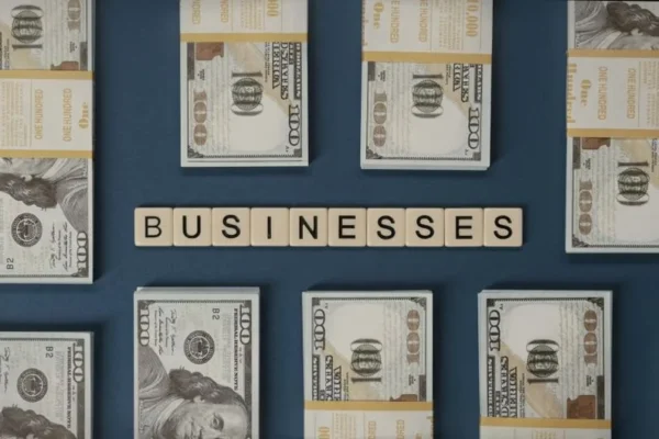 Bundles of US hundred-dollar bills and Scrabble tiles spelling 'businesses' laid out creatively.