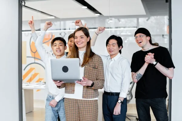 Group of diverse colleagues celebrating success while collaborating on a laptop indoors.